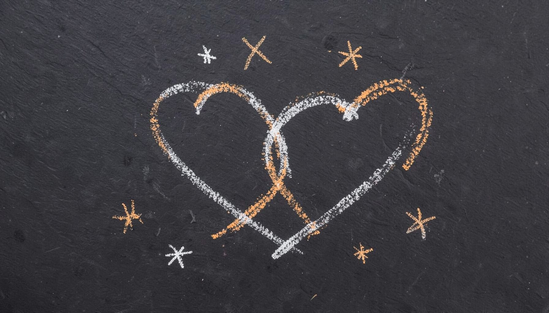 A chalk drawing on a dark slate board showing two simple interlocking heart outlines surrounded by scattered small stars in white chalk