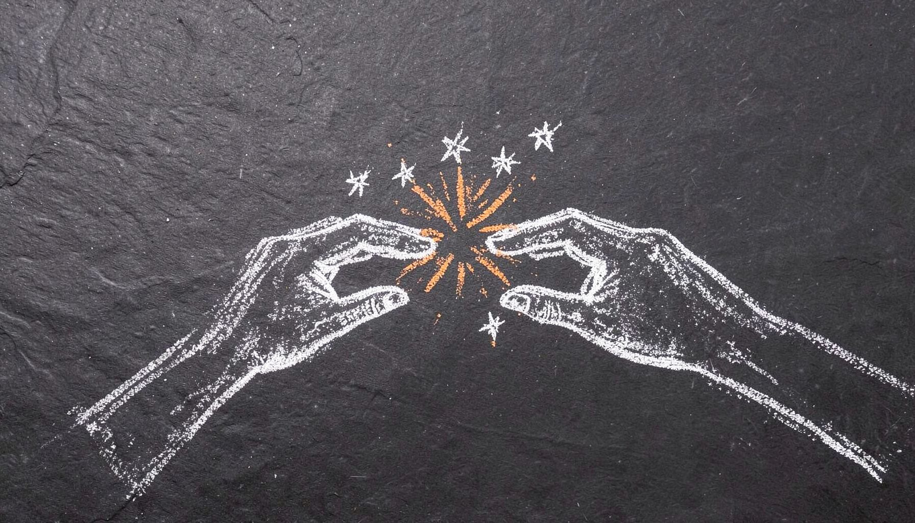 A chalk drawing on a dark slate board of two hands reaching toward each other with a small burst of stars where they nearly touch, sketched in white and warm golden chalk